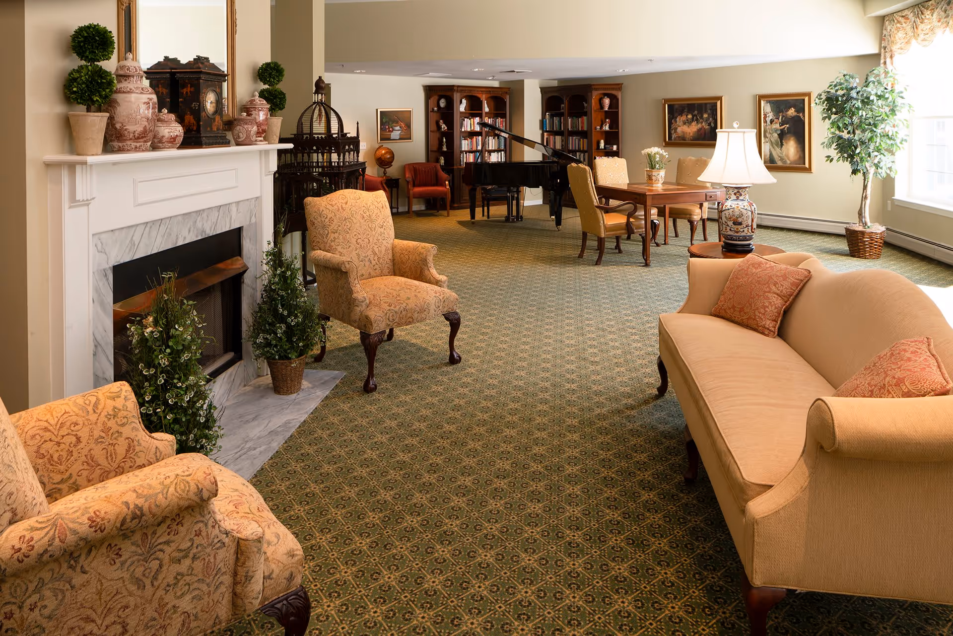 A cozy living room area with a beige sofa and two patterned armchairs arranged around a white marble fireplace decorated with vases and topiary plants. In the background, there is a grand piano, bookshelves filled with books, a wooden table with chairs, framed paintings on the walls, and a large window letting in natural light.