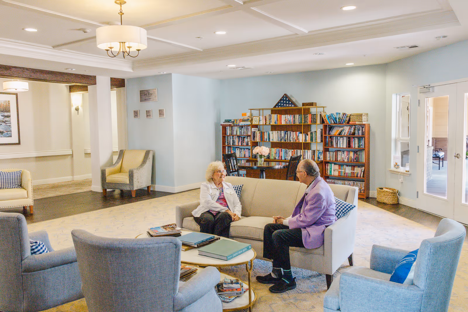 Two elderly residents sit and talk on a sofa in a bright communal living room with bookshelves and armchairs.