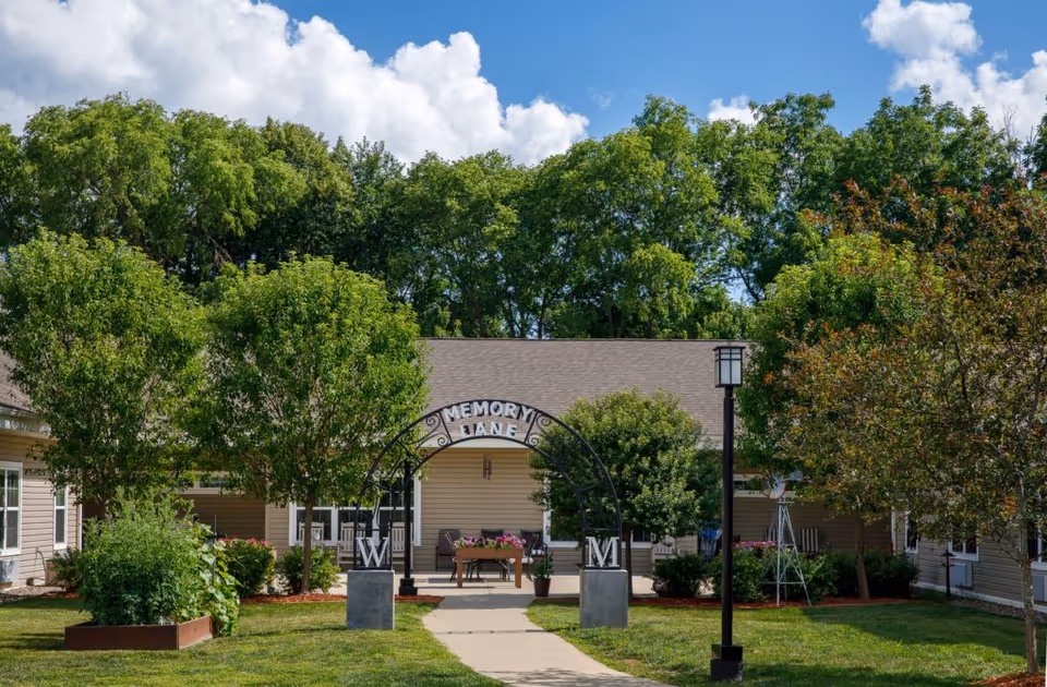 Outdoor courtyard area at Homestead Assisted Living & Memory Care of Morrison featuring a paved walkway leading through an archway labeled 'Memory Lane' with the letters W and M on stone bases. The courtyard is surrounded by green trees, shrubs, and a beige building with a porch and seating. A lamp post stands near the walkway under a partly cloudy sky.