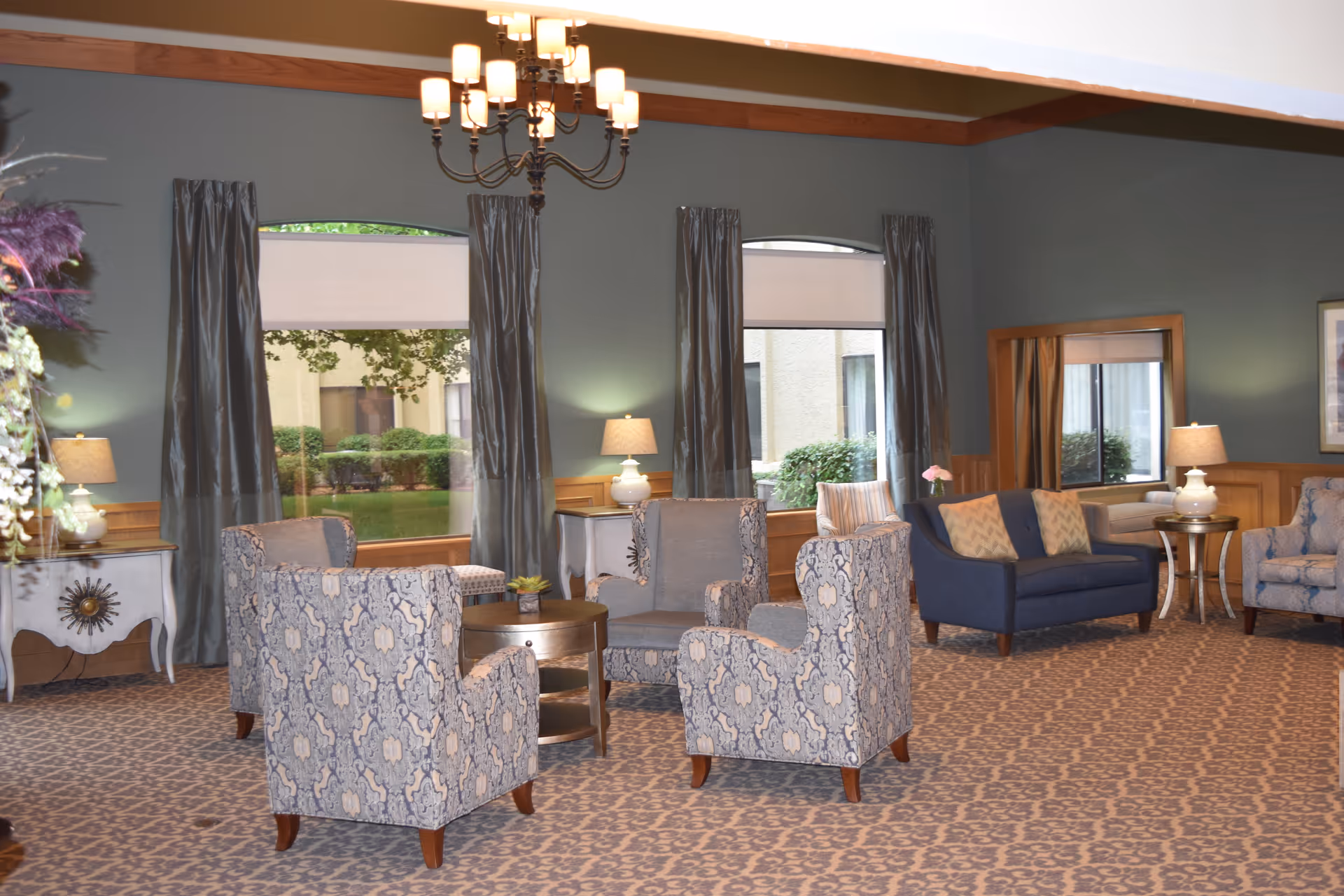 A cozy living room area with patterned armchairs arranged around a small round wooden table. There is a blue loveseat with two pillows, side tables with lamps, and large windows with gray curtains letting in natural light. The room has a patterned carpet and a chandelier hanging from the ceiling.
