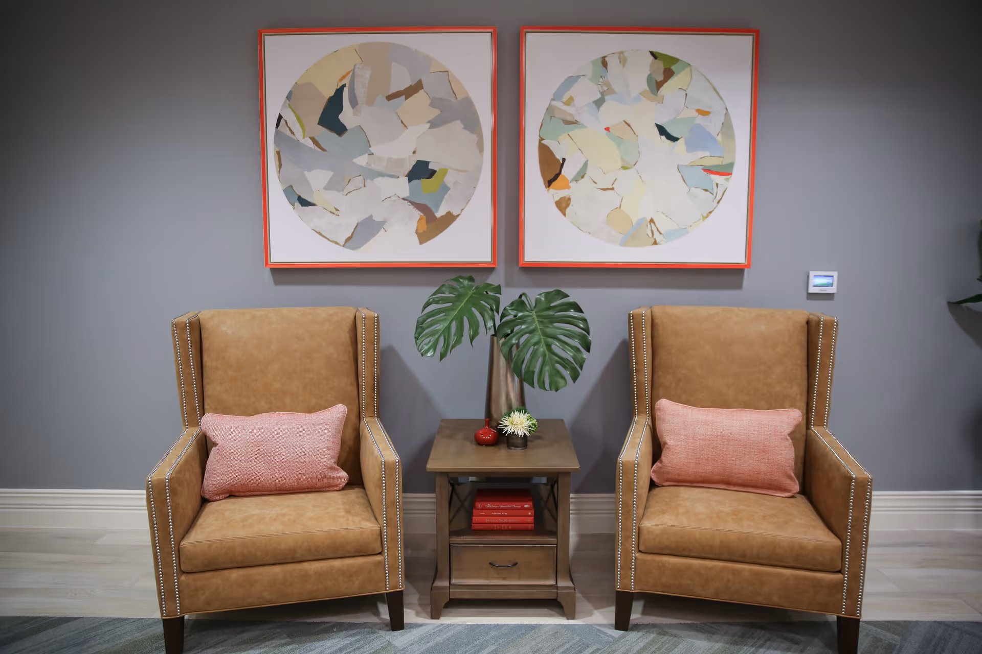 Two tan leather armchairs with pink cushions are positioned on either side of a wooden side table. The table holds a vase with large green leaves and small decorative items. Above the chairs, two abstract circular artworks with orange frames hang on a gray wall.