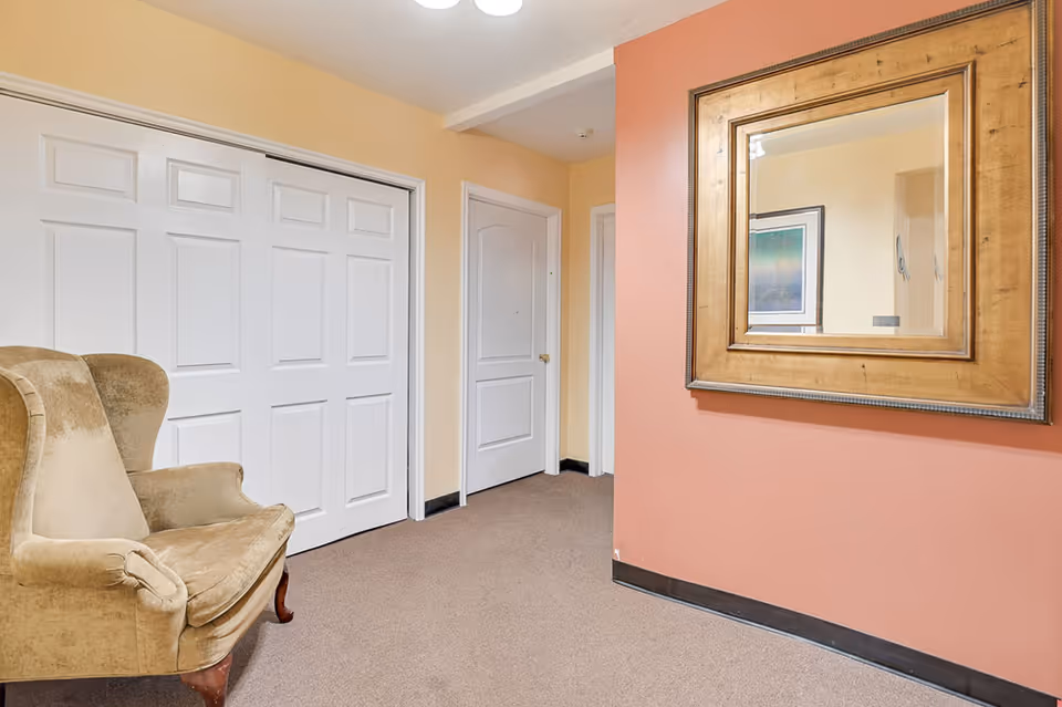 A hallway area with beige walls and carpeted floor. There is a large beige upholstered armchair on the left side, white double closet doors, and a white door further down the hallway. On the right wall, which is painted a muted coral color, there is a large square mirror with a wooden frame.