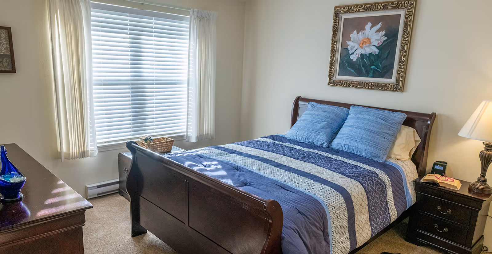 A cozy bedroom with a wooden bed frame, blue and white striped bedding, two blue pillows, a nightstand with a lamp, a book, and glasses. There is a window with white blinds and curtains, a dresser with a blue decorative vase, and a framed floral painting on the wall above the bed.