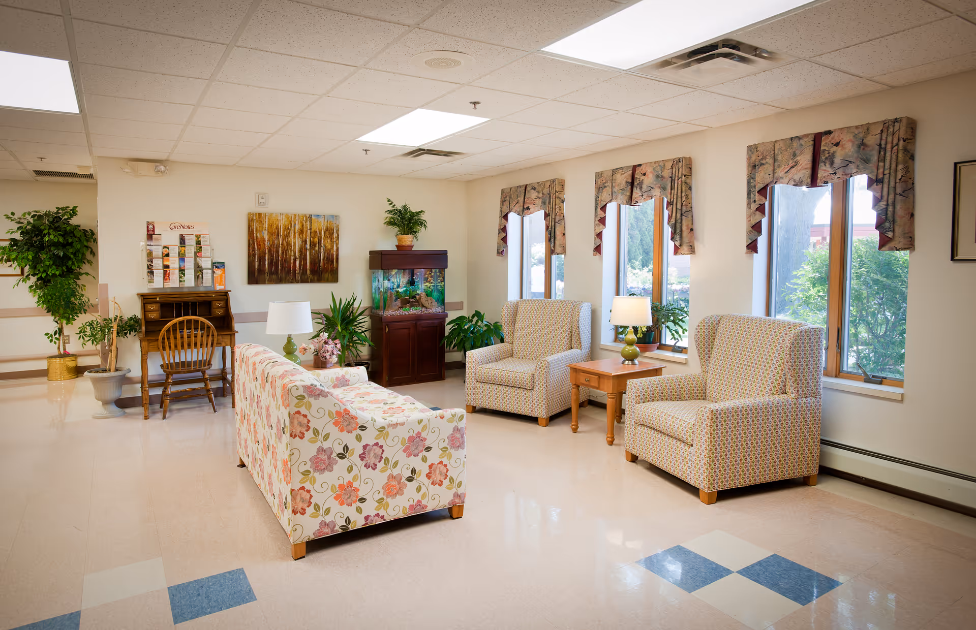 A bright communal sitting room with a floral sofa, patterned armchairs, side tables, plants, an aquarium, and windows.