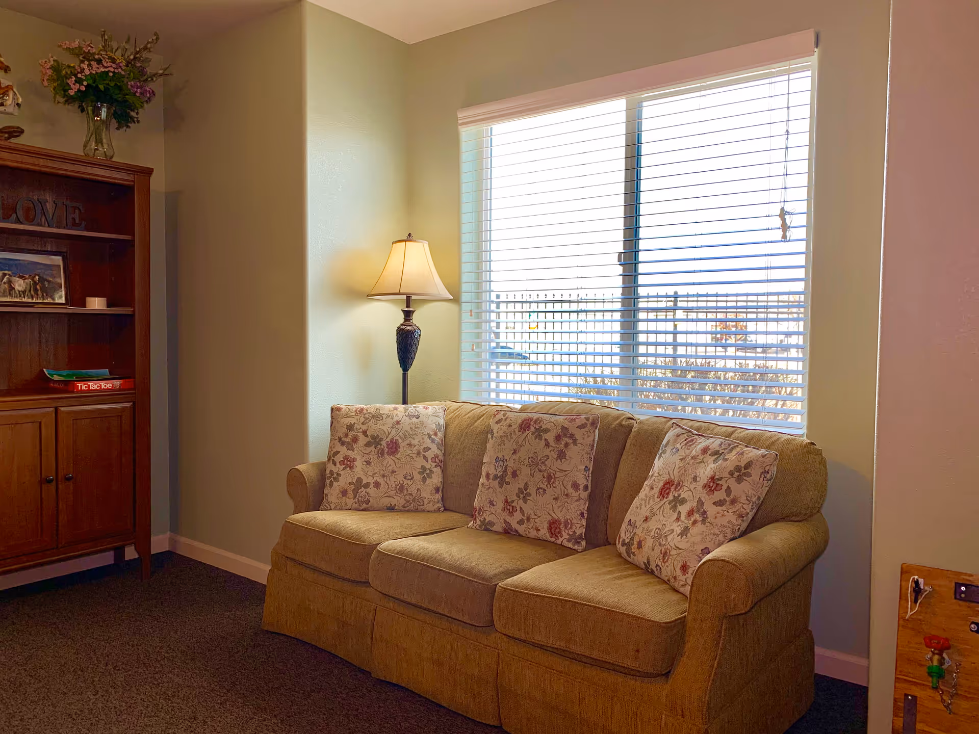 A cozy living room corner with a beige sofa adorned with three floral-patterned cushions. Next to the sofa is a floor lamp with a beige lampshade, and behind it is a large window with white blinds partially open, letting in natural light. To the left, there is a wooden cabinet with shelves holding a vase of flowers, a framed picture, and a Tic Tac Toe game. The walls are painted light green.