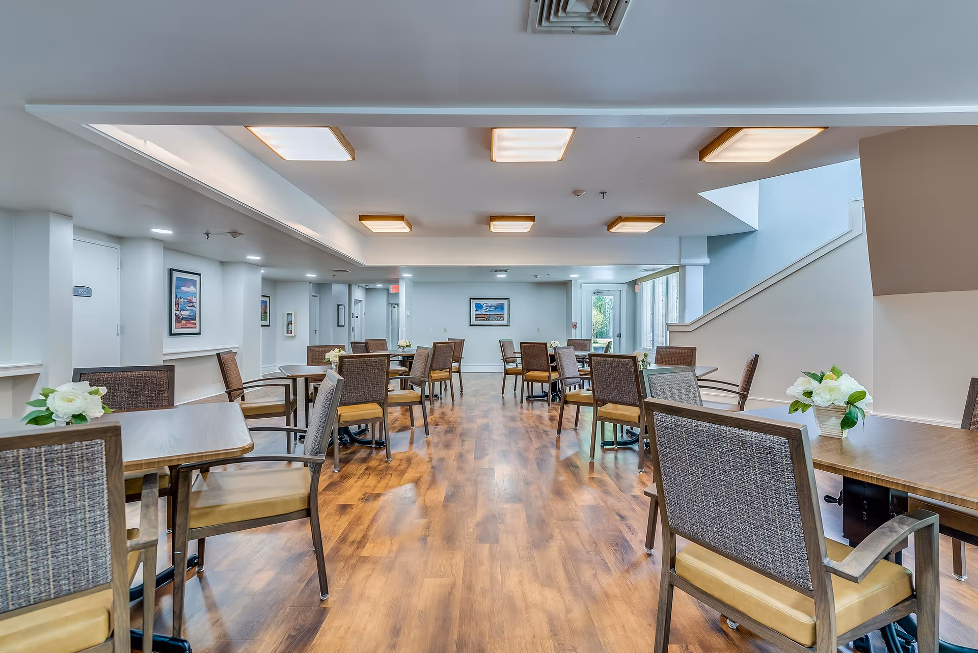 A bright and spacious dining room with multiple wooden tables and cushioned chairs arranged neatly. The floor is wooden, and the ceiling has modern square light fixtures. There are framed pictures on the walls and a door with windows letting in natural light. Small flower arrangements are placed on the tables.