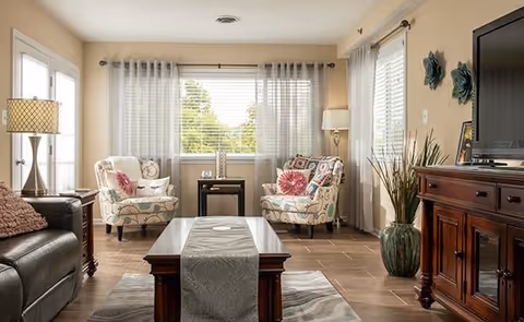 A bright and cozy living room with two patterned armchairs featuring pink and blue floral cushions, a dark leather sofa with a pink pillow, a wooden coffee table with a gray runner, a large window with sheer white curtains letting in natural light, a wooden TV cabinet with a flat-screen TV, a tall green plant in a decorative pot, and a floor lamp in the corner.