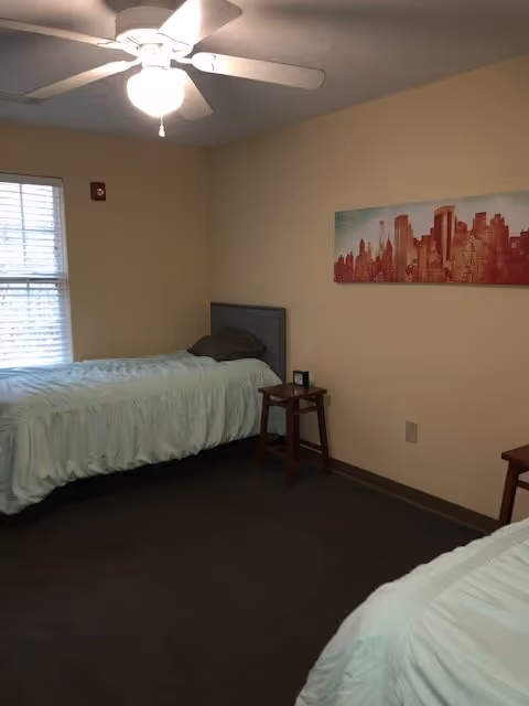 A simple bedroom with two twin beds covered in light blue bedding. There is a small wooden stool between the beds with a clock on it. A ceiling fan with a light is on, and a window with blinds is on the left wall. A red cityscape artwork hangs on the right wall.