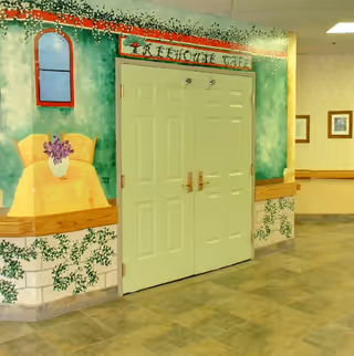 Double doors labeled 'Beehouse Cafe' set in a hallway with a mural painted on the walls depicting a window, a chair with a vase of flowers, and greenery. The floor is tiled and there are framed pictures on the adjacent wall.