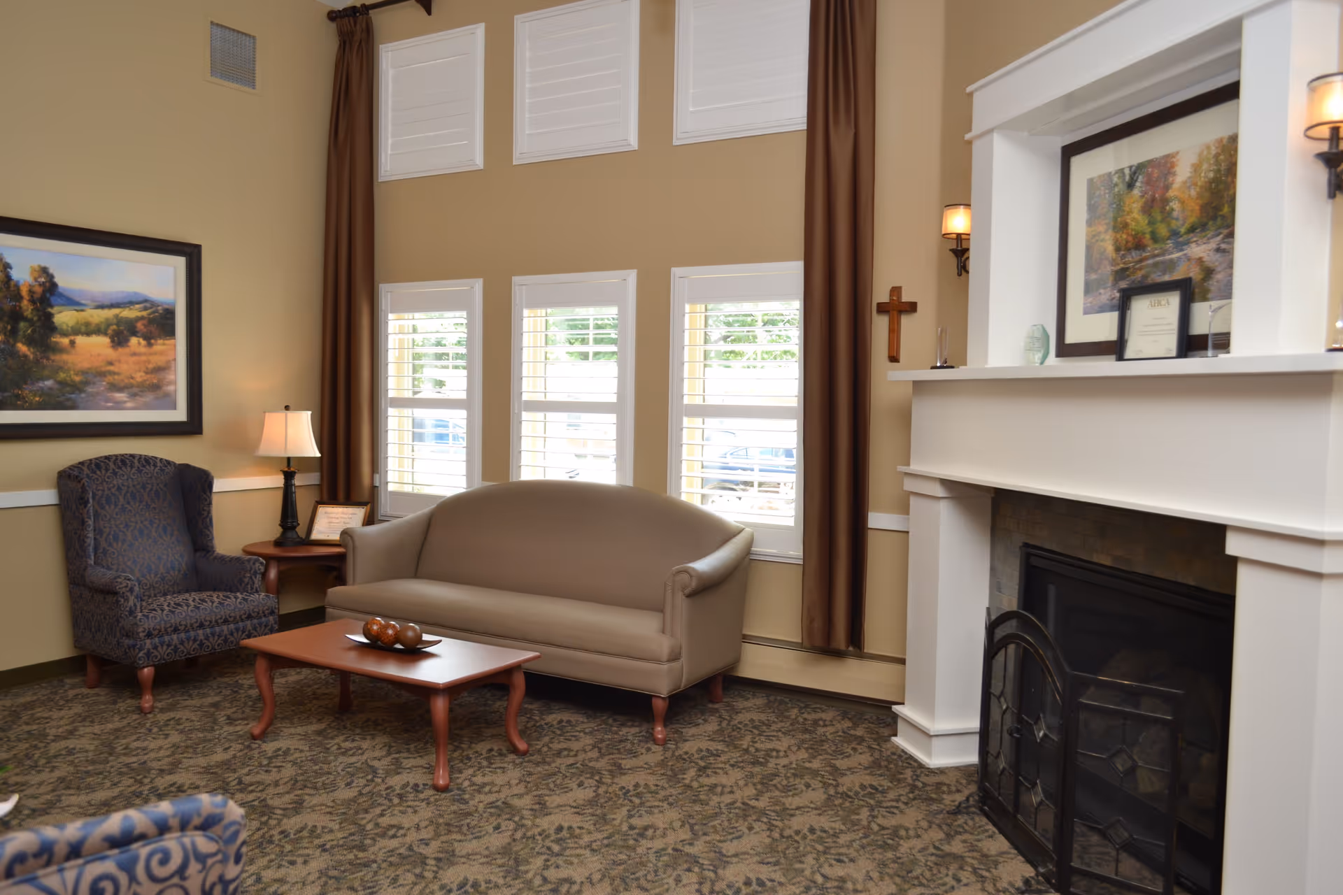 A cozy living room area with a beige sofa, a patterned armchair, a wooden coffee table with decorative items, a side table with a lamp, a fireplace with a white mantle, framed artwork on the walls, and tall windows with white shutters and brown curtains.