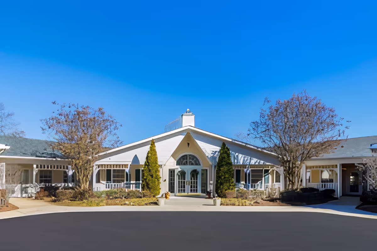 Front entrance of a single-story white senior living facility with an arched central doorway, landscaping, and a bright blue sky.