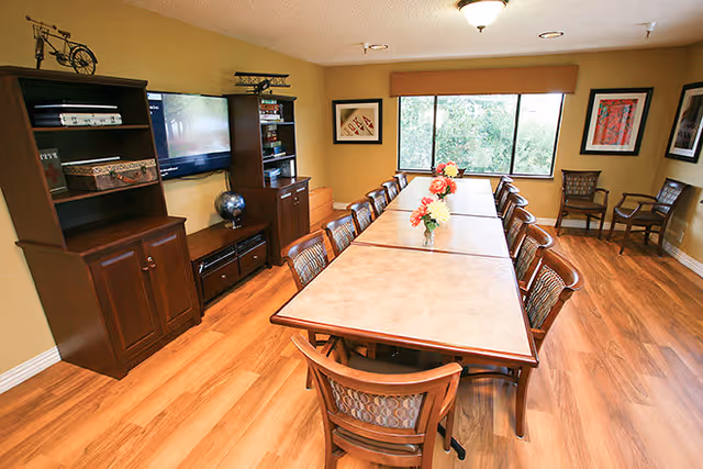 A well-lit dining room with a long rectangular table surrounded by wooden chairs. The table has three vases with flowers as centerpieces. The room features wooden flooring, a large window with a view of greenery outside, framed artwork on the walls, and a wooden cabinet with a mounted flat-screen TV.