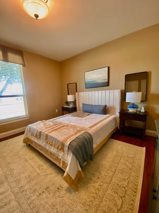 A neatly made bed with a white and beige bedspread and a gray throw blanket at the foot, positioned against a beige wall with a landscape painting above the headboard. There are two dark wooden nightstands on either side of the bed, each with a lamp and decorative items. A large window with blinds is on the left side, and a beige patterned rug covers the wooden floor.