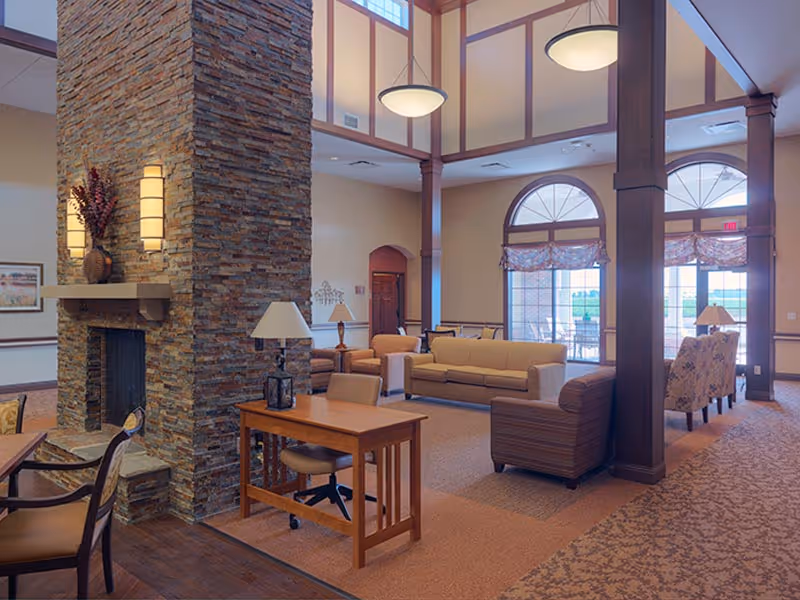 Spacious, well-lit communal lounge with a central stone fireplace, sofas, chairs, a small desk, and large arched windows.