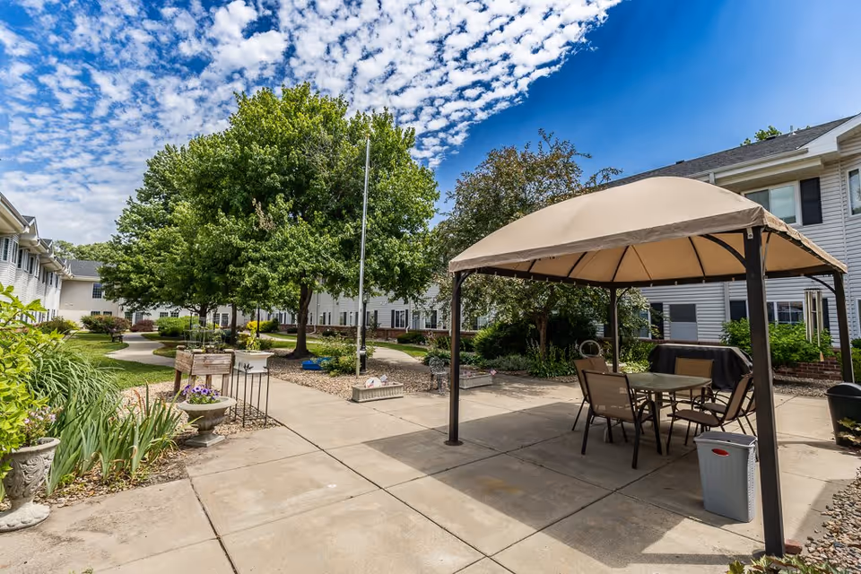 Outdoor courtyard area at Bailey Pointe Assisted Living at Van Dorn featuring a paved walkway, green trees, flower beds, and a shaded seating area with a table and chairs under a beige canopy. The building surrounds the courtyard with white siding and multiple windows under a partly cloudy blue sky.