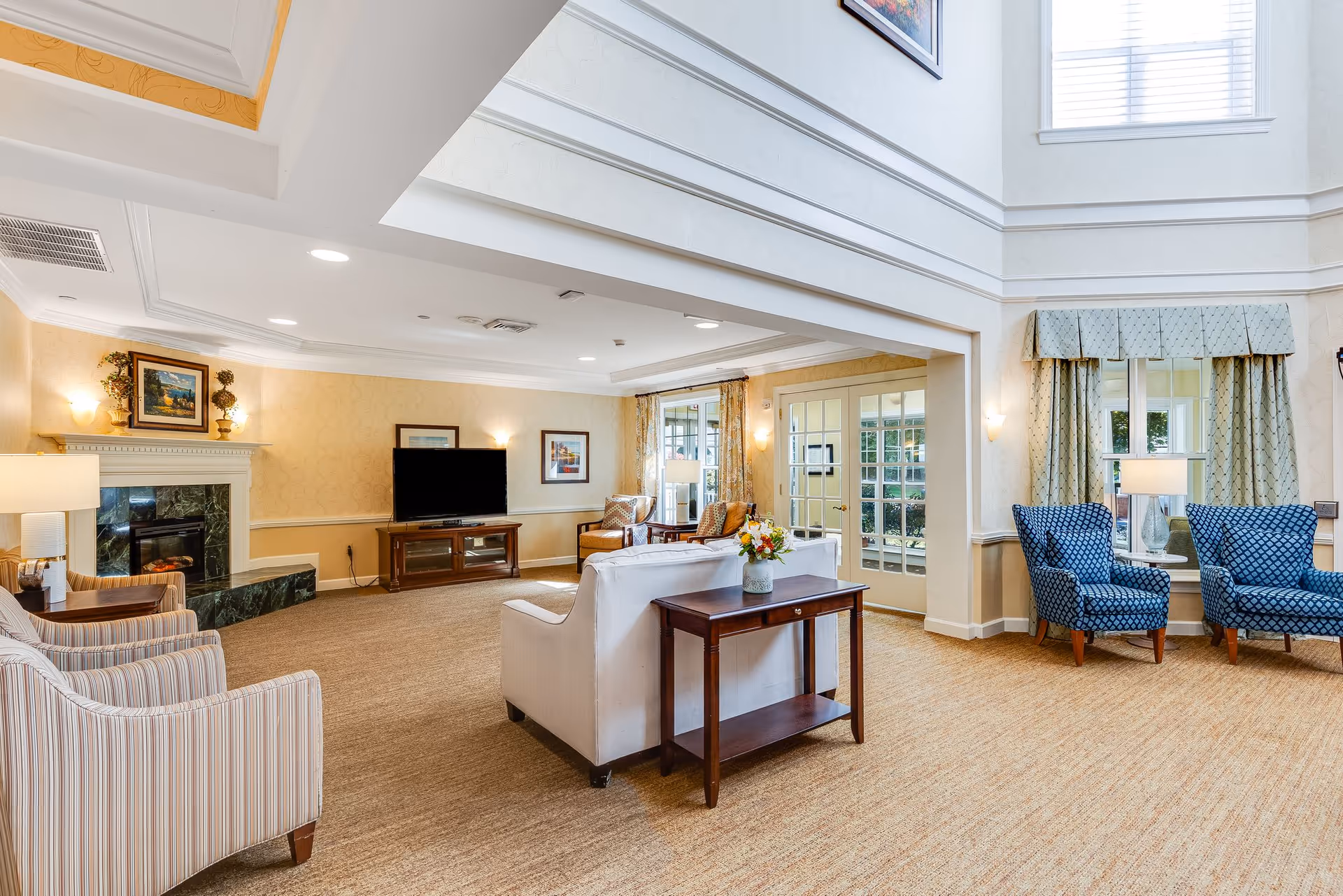 A spacious and well-lit living room area in a senior living facility featuring a white sofa with a wooden side table holding a flower vase, two striped armchairs, two blue patterned armchairs near windows with curtains, a fireplace with a painting above it, a flat-screen TV on a wooden stand, and framed artwork on the walls. The room has beige carpet and light-colored walls with crown molding and recessed lighting.
