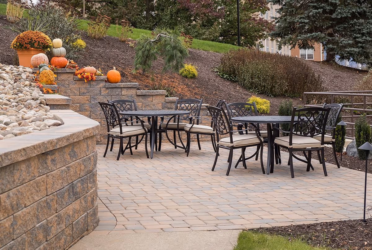 Outdoor patio area with two black metal tables and eight matching chairs with beige cushions. The patio is paved with stone tiles and surrounded by landscaped garden beds with mulch, small shrubs, and decorative pumpkins and flowers on stone steps.