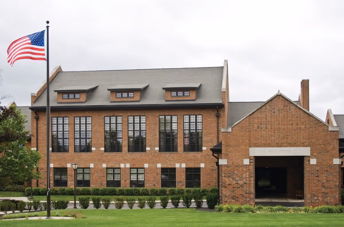 Exterior view of a large brick building with multiple tall windows and a gray roof. There is a covered entrance on the right side with the words 'Star Points' above it. An American flag on a flagpole is visible on the left side, along with green shrubs and a well-maintained lawn in the foreground under a cloudy sky.