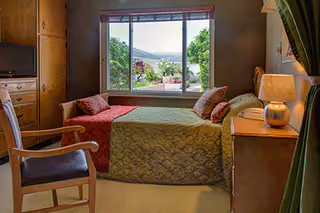 A cozy bedroom with a bed, nightstand, lamp, dresser, and window view.