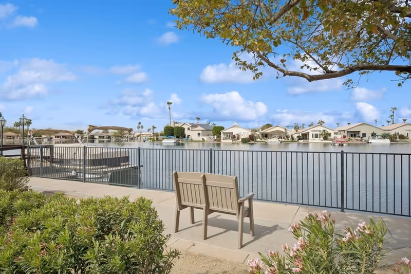 A peaceful outdoor scene at The Forum at Desert Harbor showing a bench facing a calm body of water with houses and boats on the opposite shore under a blue sky with scattered clouds. There are green shrubs and flowering plants in the foreground and a metal railing along the water's edge.