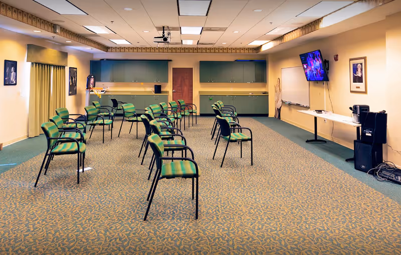 Spacious interior activity room with rows of green chairs facing a wall-mounted TV, cabinets, and a projector.