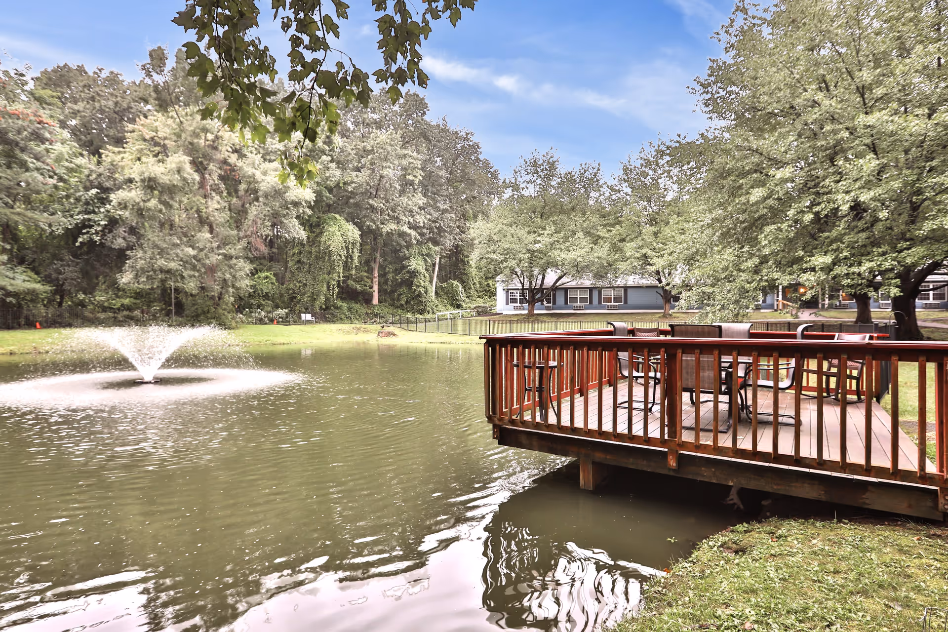 A peaceful outdoor scene at Peregrine Senior Living at Crossgate featuring a wooden deck with chairs and tables extending over a pond with a water fountain. The background includes lush green trees and a building partially visible behind the trees.