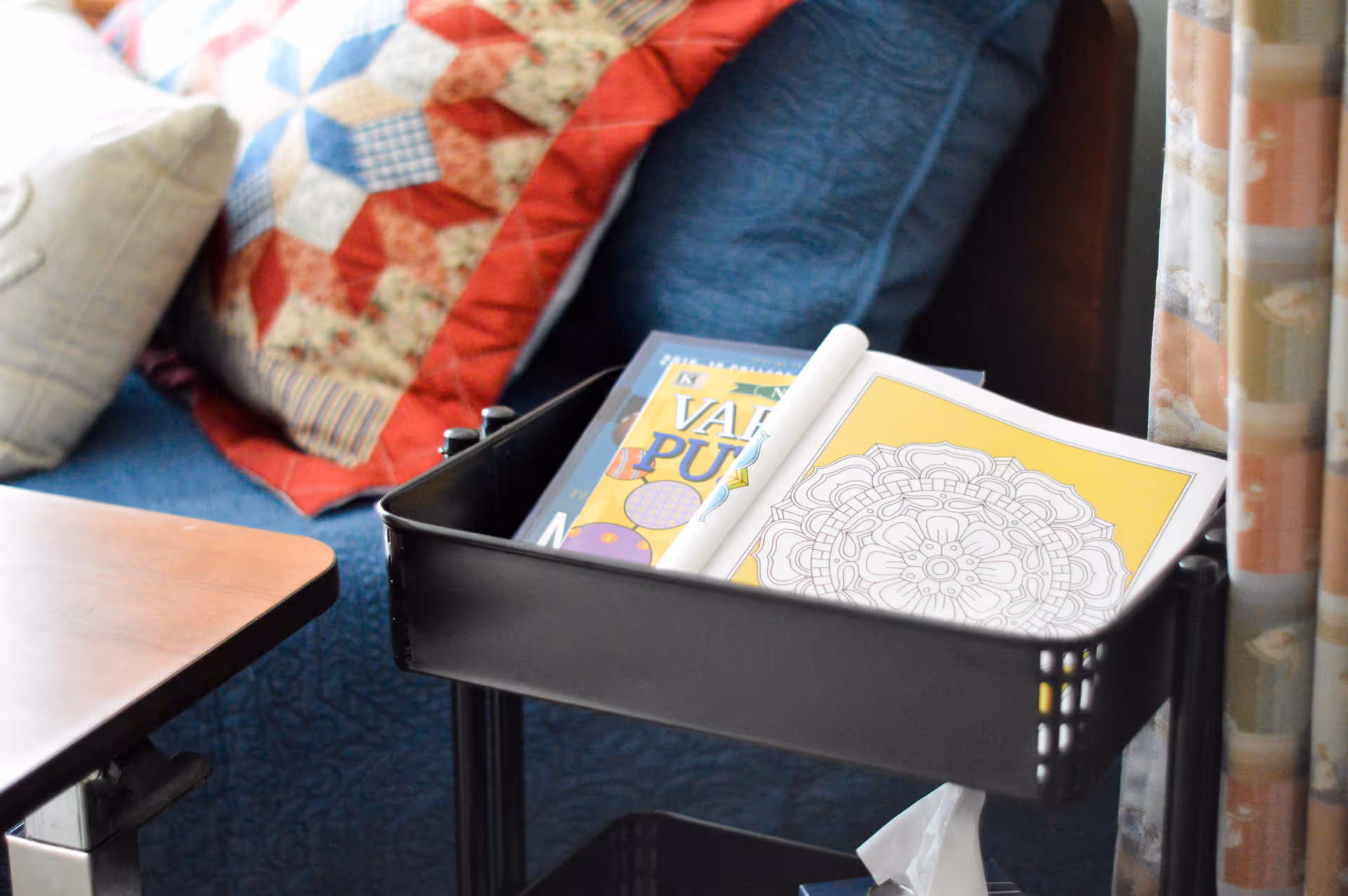 A close-up view of a black rolling cart holding a coloring book with a mandala design and a puzzle book, placed next to a blue couch with a red and beige patchwork quilt and pillows.