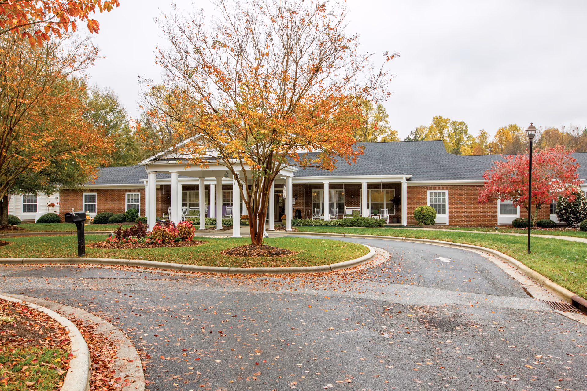 Front entrance of a single-story brick senior living building with white columns, a covered porch with rocking chairs, and a circular driveway framed by autumn trees.