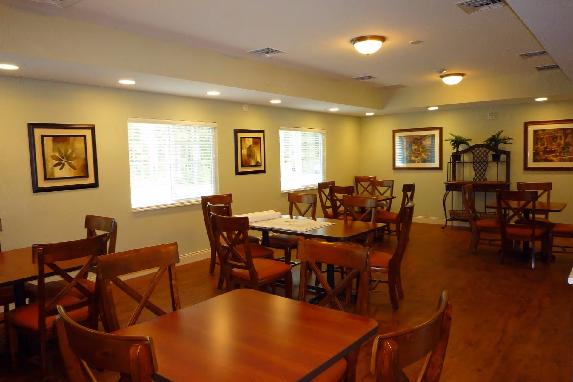 A dining room with multiple wooden tables and chairs arranged neatly. The room has light-colored walls adorned with framed artwork, windows with blinds allowing natural light, and ceiling lights providing warm illumination. There is a decorative metal and wood shelving unit with plants in the corner.