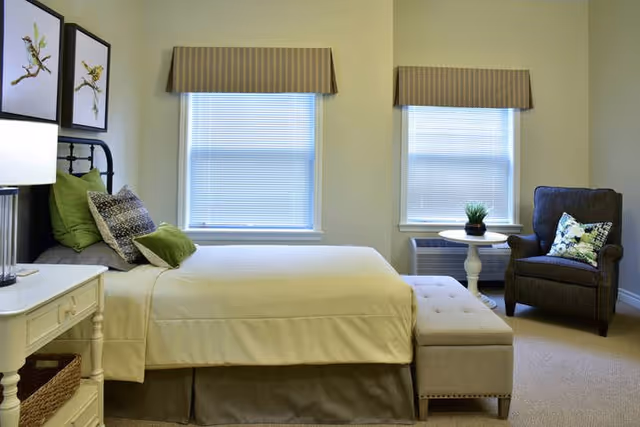 A bright, neatly furnished bedroom with a bed, ottoman at the foot, an armchair and side table by two windows.