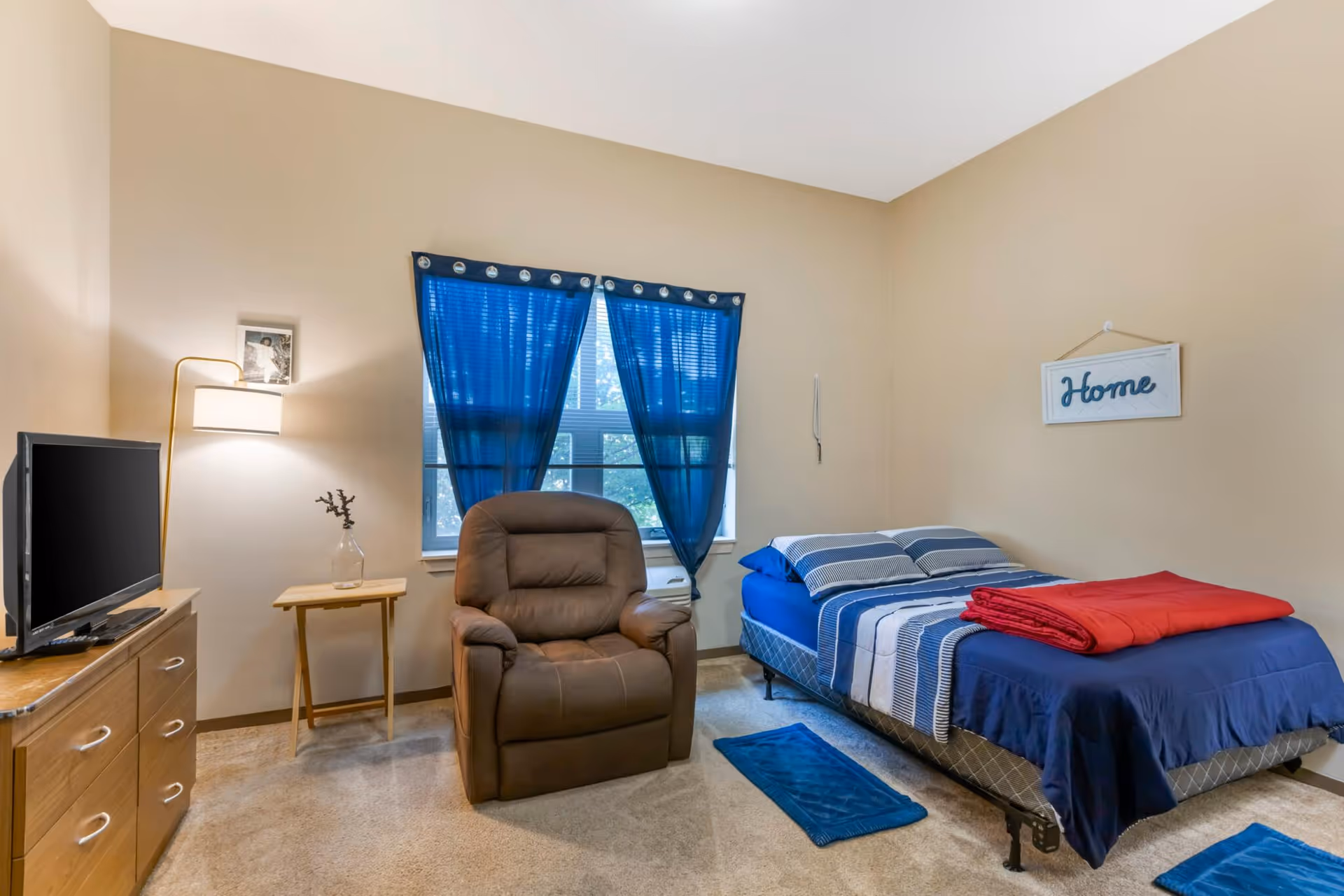 A cozy bedroom with beige walls featuring a single bed with blue and white striped bedding and a red blanket folded on top. There is a brown recliner chair in front of a window with blue curtains. A wooden dresser with a flat-screen TV and a small side table with a vase and a lamp are also visible. A wall decoration with the word 'Home' hangs above the bed.