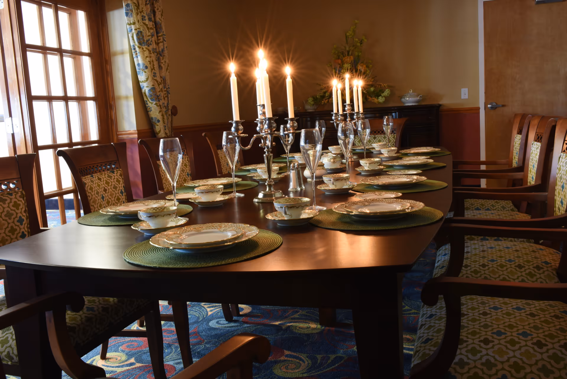 A formal dining room with a long wooden table set for a meal. The table has green placemats, fine china plates, teacups, and tall champagne glasses. Two silver candelabras with lit candles are placed along the center of the table. The room has patterned upholstered chairs, floral curtains, a wooden sideboard with a floral arrangement, and a closed wooden door.