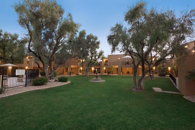 A spacious outdoor courtyard area at Scottsdale Village Square with green grass, several mature trees, a central water fountain, and surrounding two-story buildings with balconies and outdoor seating. The sky is clear and the area is softly lit with outdoor lights.