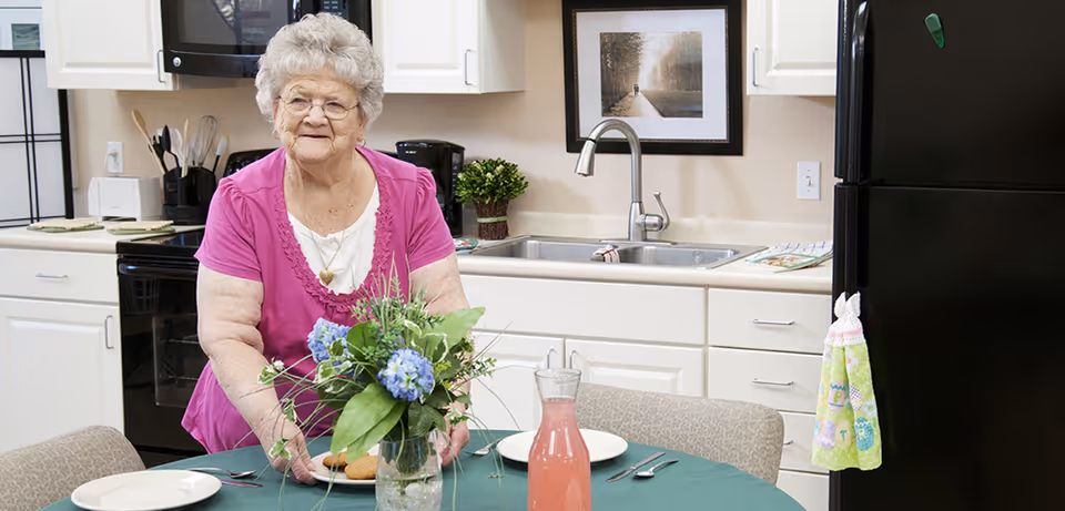 A person stands in a bright kitchen-dining area arranging a vase of flowers on a table set with plates and a pitcher.