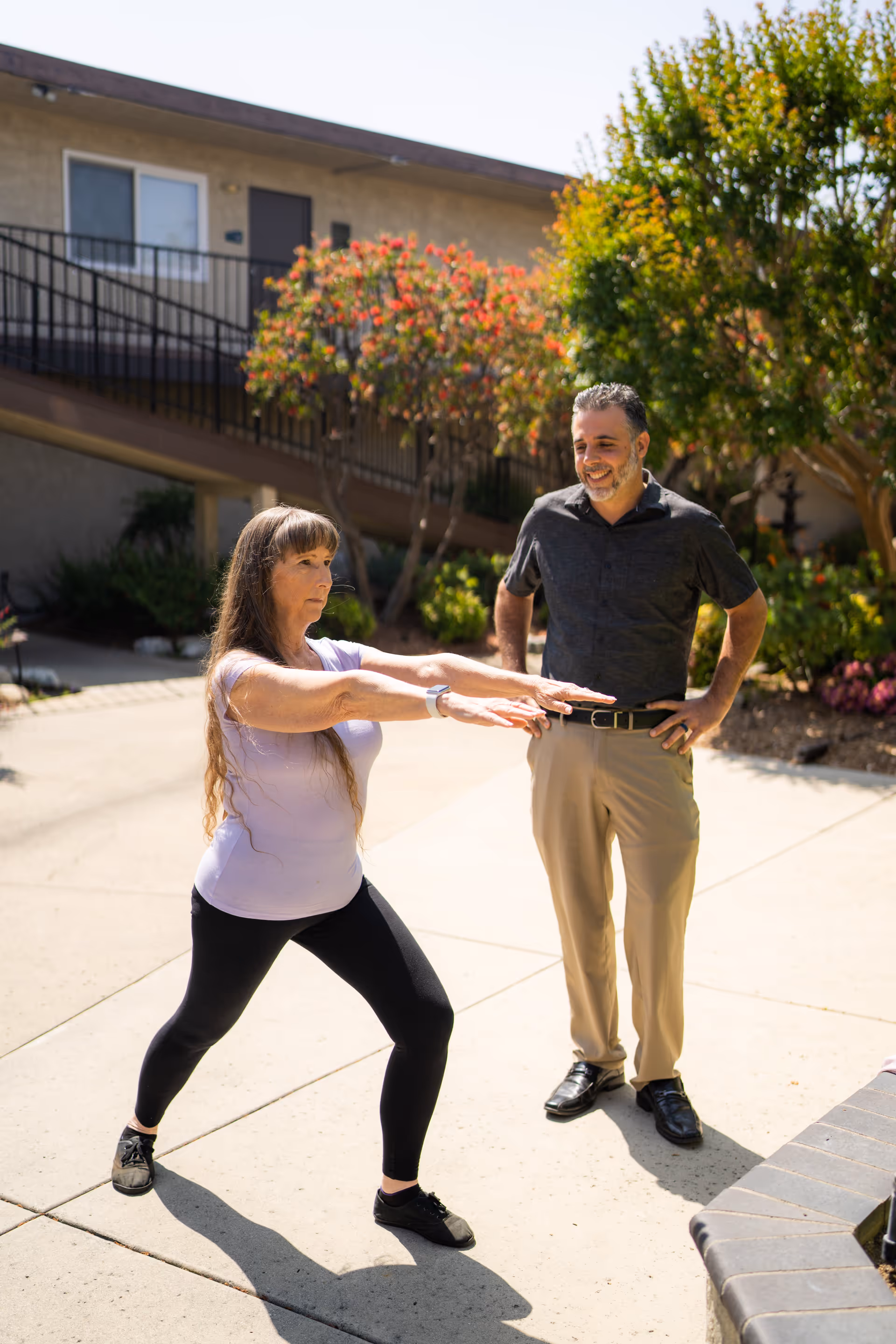A woman in black leggings and a white shirt is performing a stretching exercise outdoors on a paved area, while a man in a dark shirt and beige pants watches and smiles. They are in front of a building with stairs and surrounded by greenery and flowering bushes.