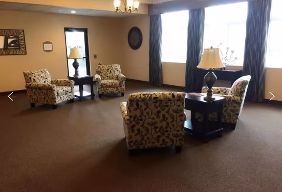 A sitting area in a senior living facility with four patterned armchairs arranged around two dark wooden side tables, each with a decorative lamp. Large windows with dark curtains allow natural light to fill the room, and a chandelier hangs from the ceiling. The walls are decorated with framed artwork and a circular wall piece.