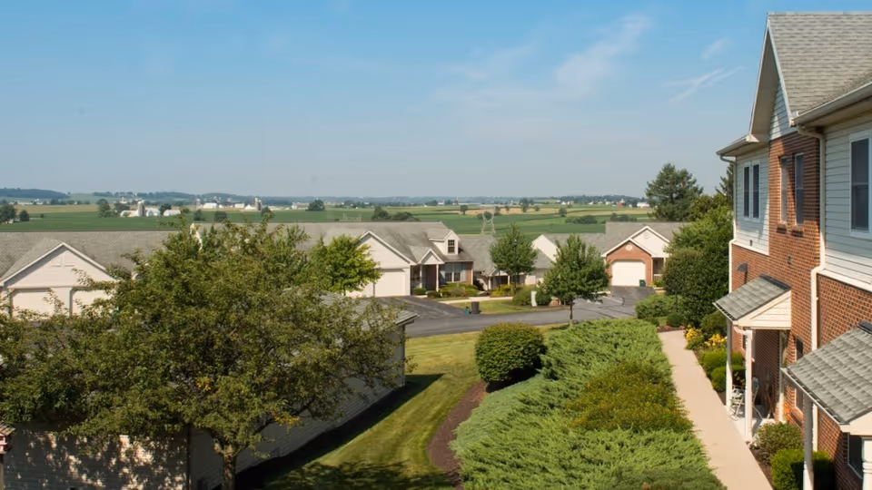 View of the retirement community showing brick residential buildings, a landscaped walkway, shrubs and trees with open fields under a blue sky.