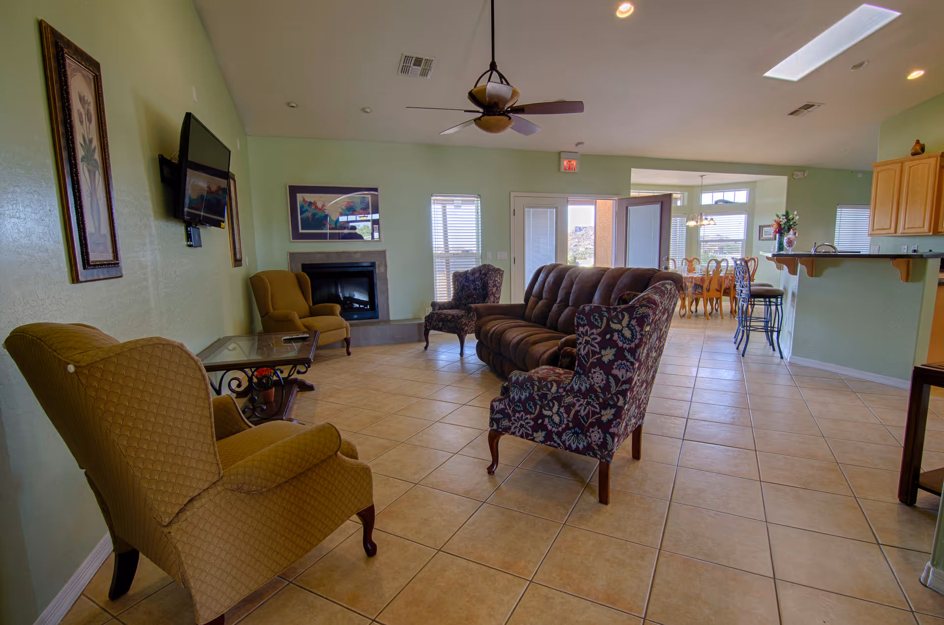 A spacious living room with tiled floors, light green walls, and a ceiling fan. The room features a brown sofa, two patterned armchairs, two yellow armchairs, a glass-top side table, a wall-mounted TV, and a fireplace. In the background, there is a dining area with a wooden dining table and chairs, and a kitchen counter with bar stools.