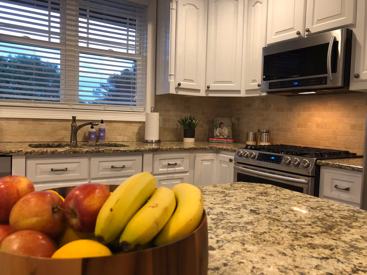 A modern kitchen with white cabinets, granite countertops, a stainless steel microwave and stove, a sink under a window with blinds, and a bowl of fresh fruit including bananas, apples, and a lemon on the counter.