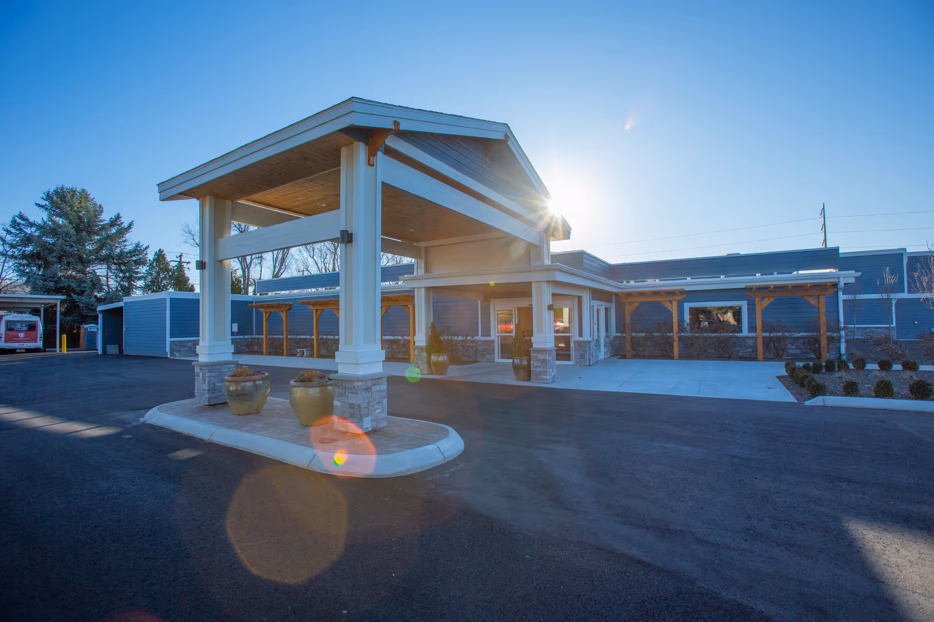 Exterior view of Heatherwood Senior Living facility entrance with a covered drop-off area, potted plants, and a clear blue sky with the sun shining brightly.