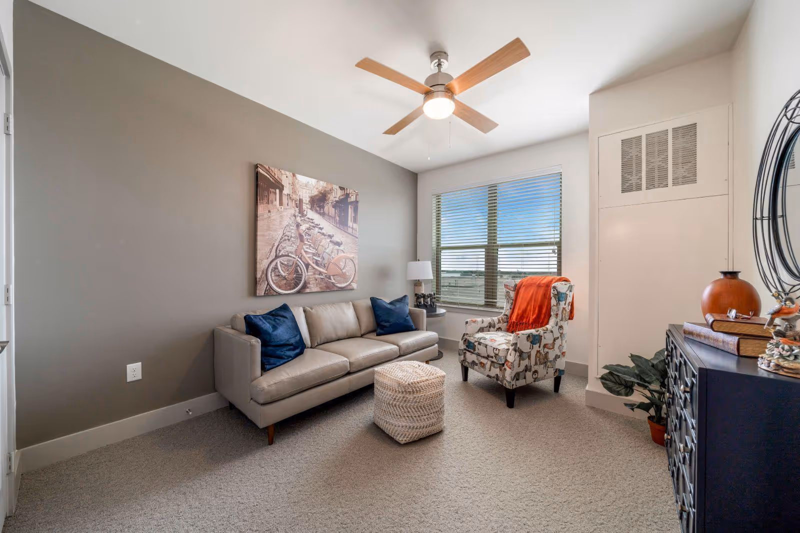 A cozy living room with a beige sofa adorned with two dark blue pillows, a patterned armchair with an orange throw blanket, a woven ottoman, a ceiling fan with wooden blades, a large window with blinds, a side table with a lamp, a black dresser with decorative items, and a wall art featuring bicycles on a street.