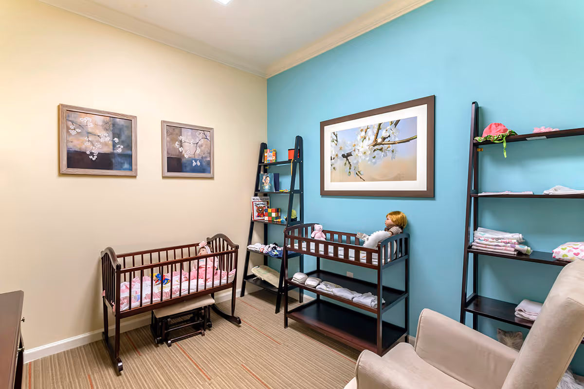 A pastel-colored nursery room with two wooden cribs, shelving stocked with toys and folded linens, and a beige armchair.
