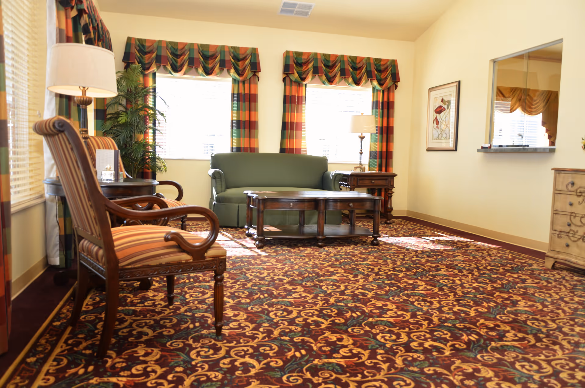 Sunlit sitting room with patterned carpet, striped armchairs, a green sofa, coffee table, and colorful window valances.