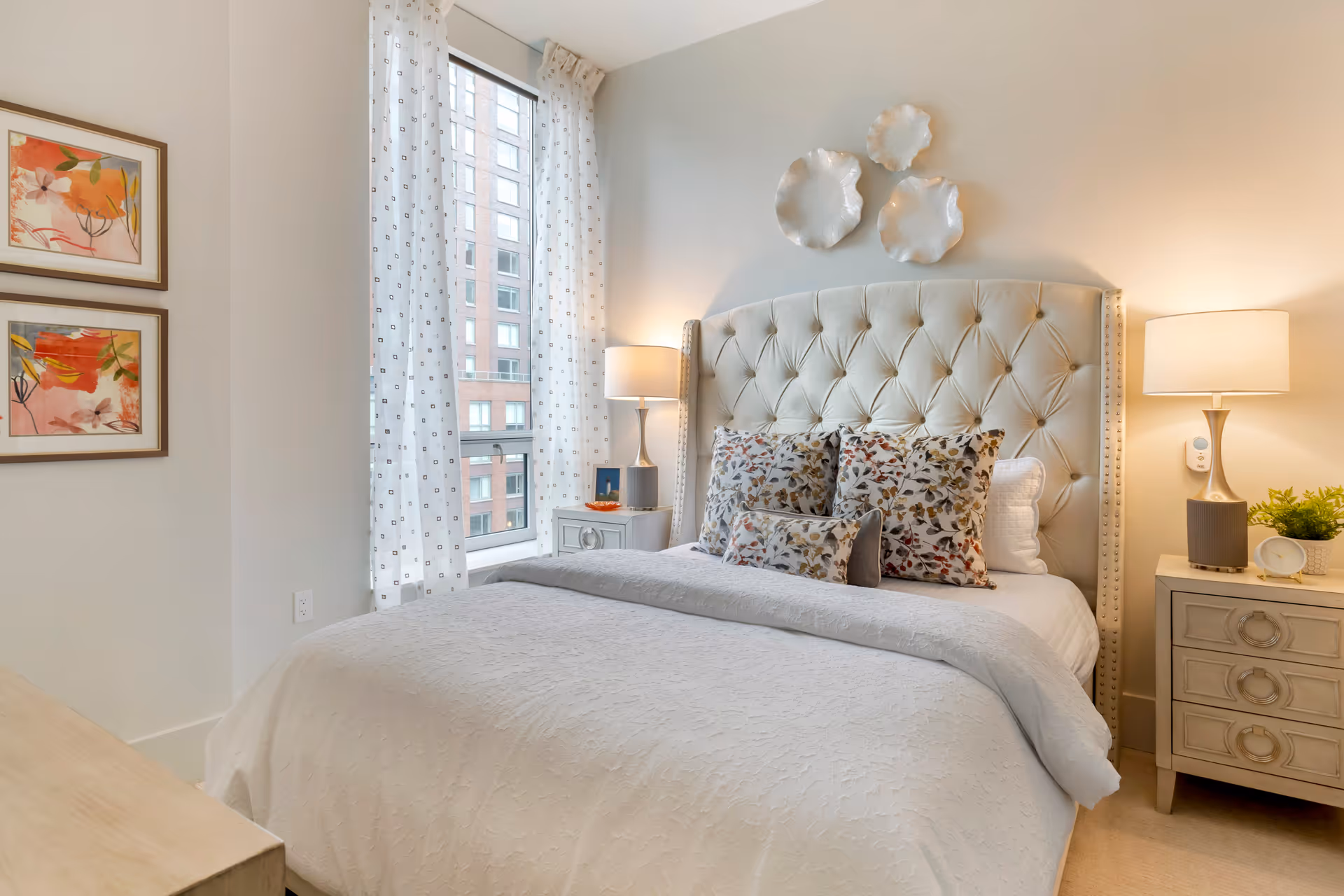A cozy bedroom with a tufted beige headboard, white bedding, and decorative floral pillows. Two bedside tables each hold a lamp, with one also featuring a small plant and clock. There are three white decorative wall pieces above the headboard, two framed floral paintings on the left wall, and a window with sheer white curtains showing a view of a brick building outside.