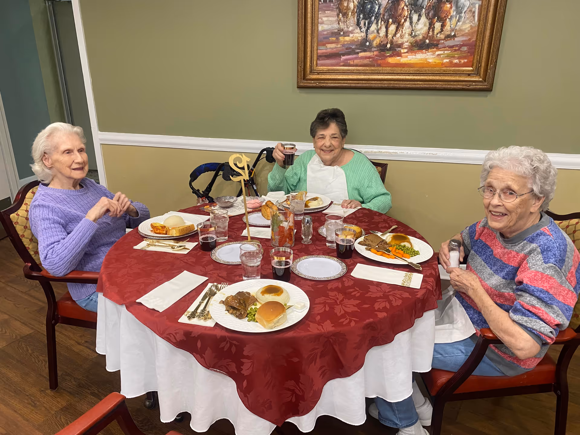 Three elderly women sitting around a round dining table with a red and white tablecloth, enjoying a meal together. The table is set with plates of food including meat, peas, carrots, rolls, and drinks. One woman is holding a glass and smiling, another is holding a salt shaker, and the third is sitting with her hands clasped. A painting of horses is hanging on the green wall behind them.
