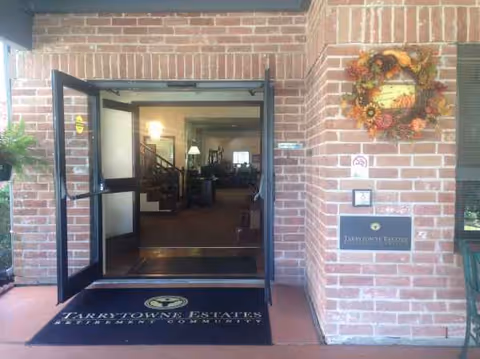 Brick building entrance with open glass doors, a wreath on the wall, and a welcome mat.