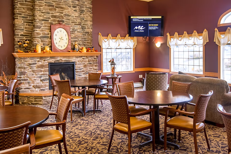 Cozy common room with a stone fireplace, round dining tables and chairs, a TV, and sofas by large windows.