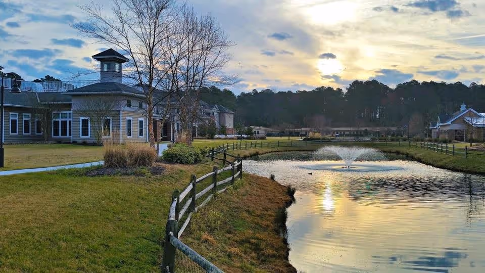 A scenic outdoor view of Warwick Forest, a CCRC, featuring a pond with a water fountain, a wooden fence along the grassy bank, leafless trees, and buildings in the background under a partly cloudy sky during sunset.