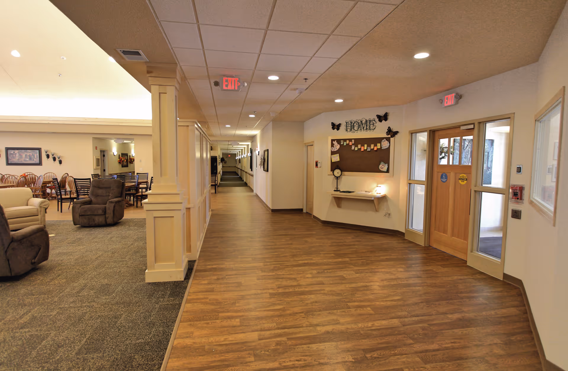Interior view of a senior living facility hallway with wood flooring and a carpeted seating area to the left featuring recliners and dining tables with chairs. On the right side, there is a bulletin board with the word 'HOME' and decorative butterflies above it, next to a set of double wooden doors with glass panels. The hallway extends into the distance with ceiling lights and exit signs visible.