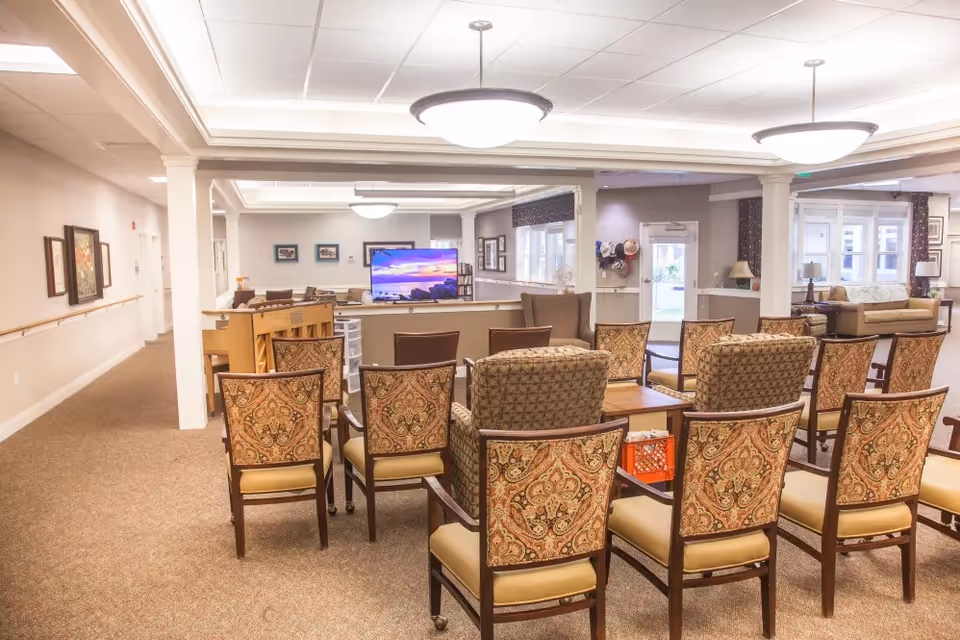 A well-lit common area in a senior living facility with multiple patterned chairs arranged in rows facing a large flat-screen TV. The room has beige walls, carpeted floors, and ceiling lights. There are couches and tables in the background, along with framed pictures on the walls and a door leading outside.